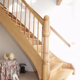 Jasne, drewniane schody z balustradą i ozdobnym słupkiem, widoczne od dołu, z dekoracjami pod schodami: stolik z kwiatowym obrusem, ozdobne jaja i stary telefon.