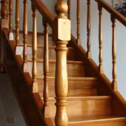 Klasyczne, drewniane schody z toczonymi balustradami i ozdobnym słupkiem z kulą na szczycie, wykończone lakierem o ciepłym, miodowym odcieniu.