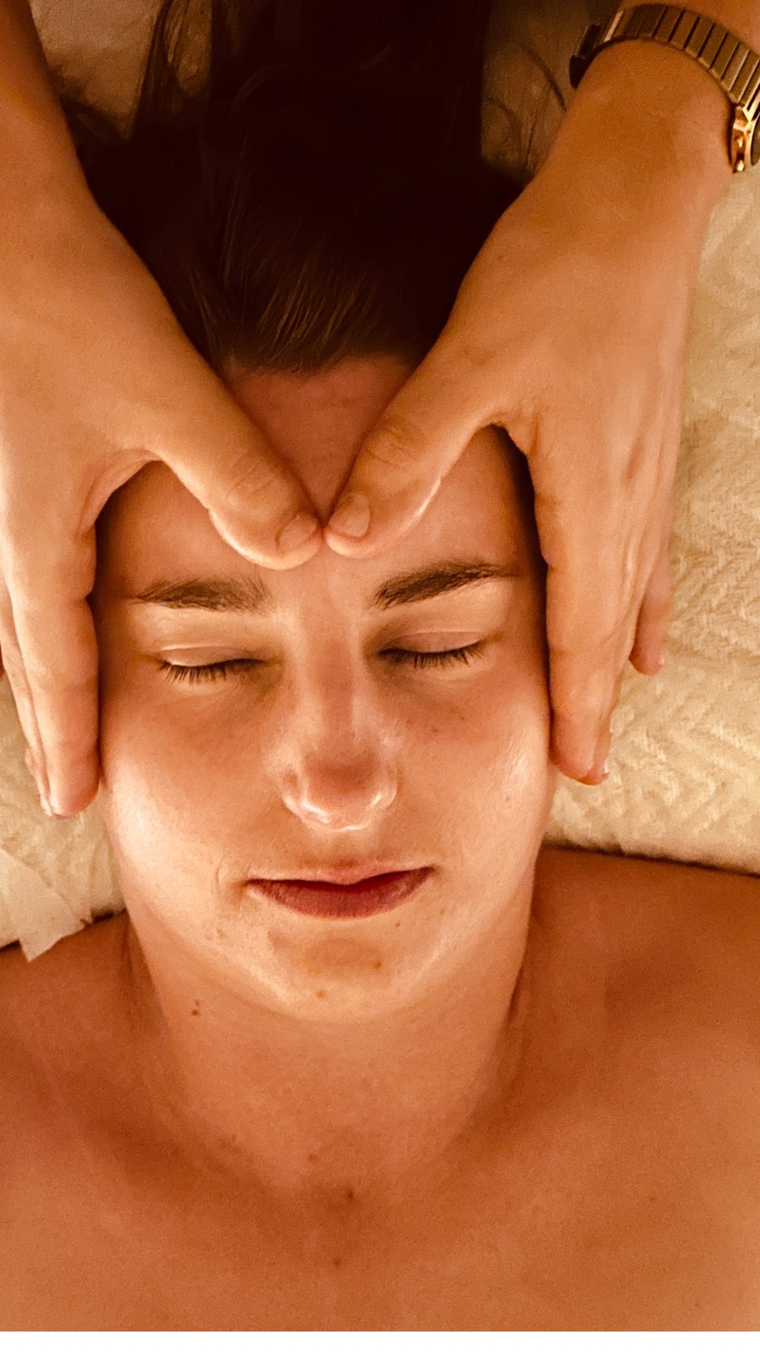 Kobieta leżąca na plecach podczas masażu twarzy. Dłonie masażysty delikatnie dotykają jej czoła, a na nadgarstku masażysty widoczny jest złoty zegarek.