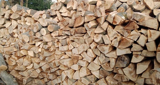 Stos pociętego drewna kominkowego ułożonego w pryzmę, widoczne sęki i naturalny kolor drewna, w tle zieleń drzew. Drewno przygotowane do sezonu grzewczego.