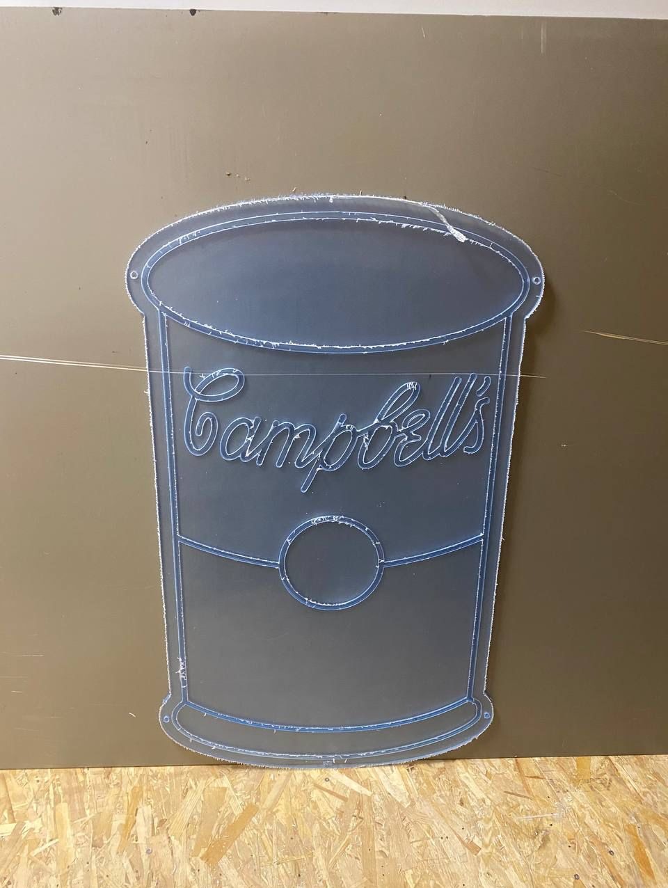 Wycięty z szarej płyty wizerunek puszki Campbell's z widocznymi śladami po frezie, na tle ściany i płyty OSB.