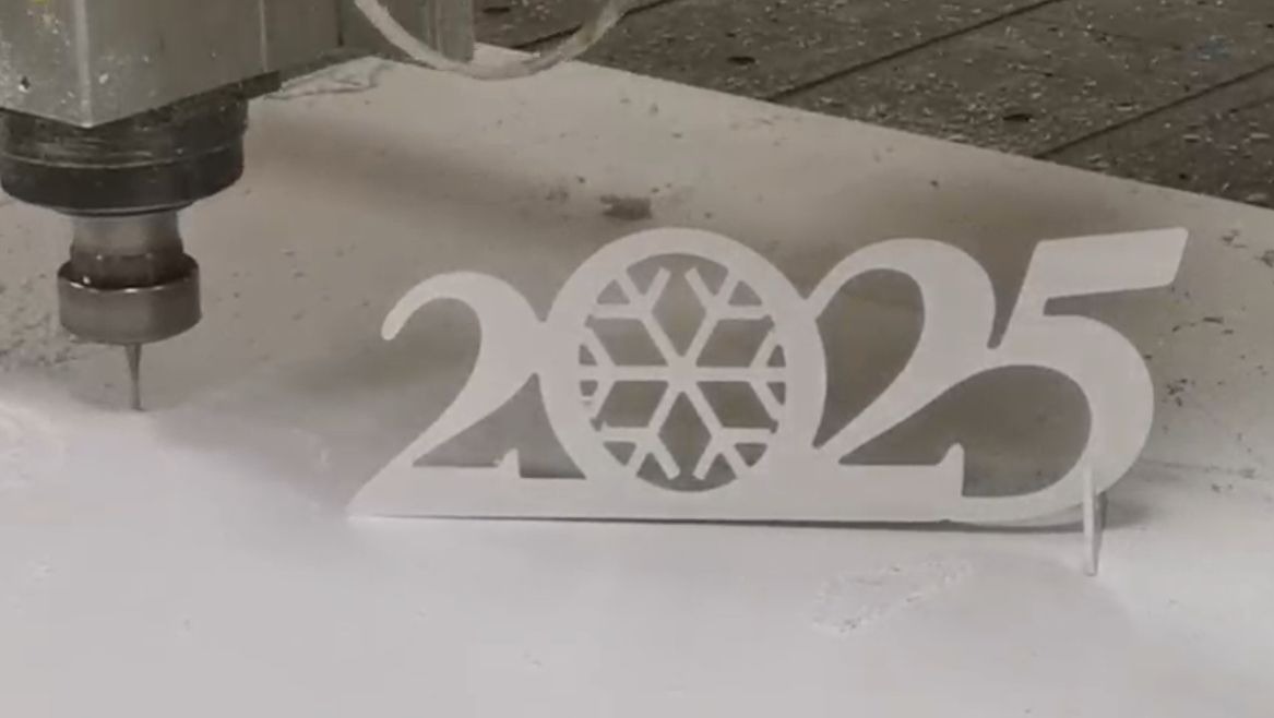 Frezarka CNC wycina z białego tworzywa sztucznego dekorację w kształcie napisu 2025 ze śnieżynką w miejscu zera.