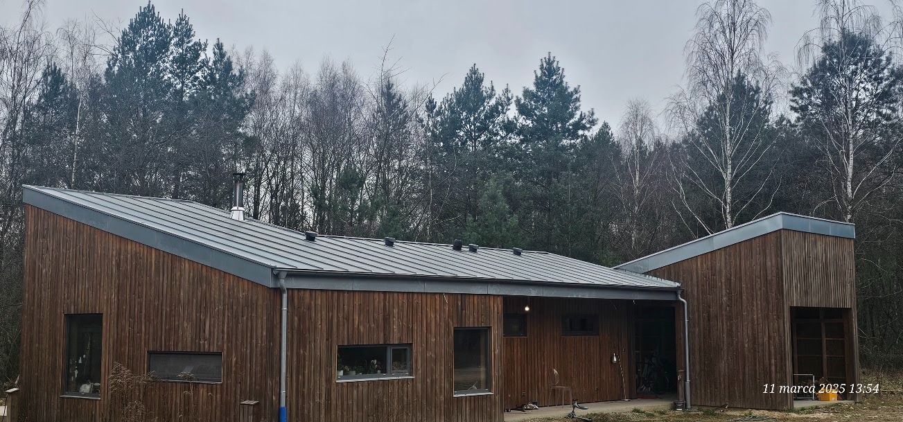 Nowoczesny dom z drewnianą elewacją i szarym, metalowym dachem na tle lasu, widoczne kominy wentylacyjne na dachu.