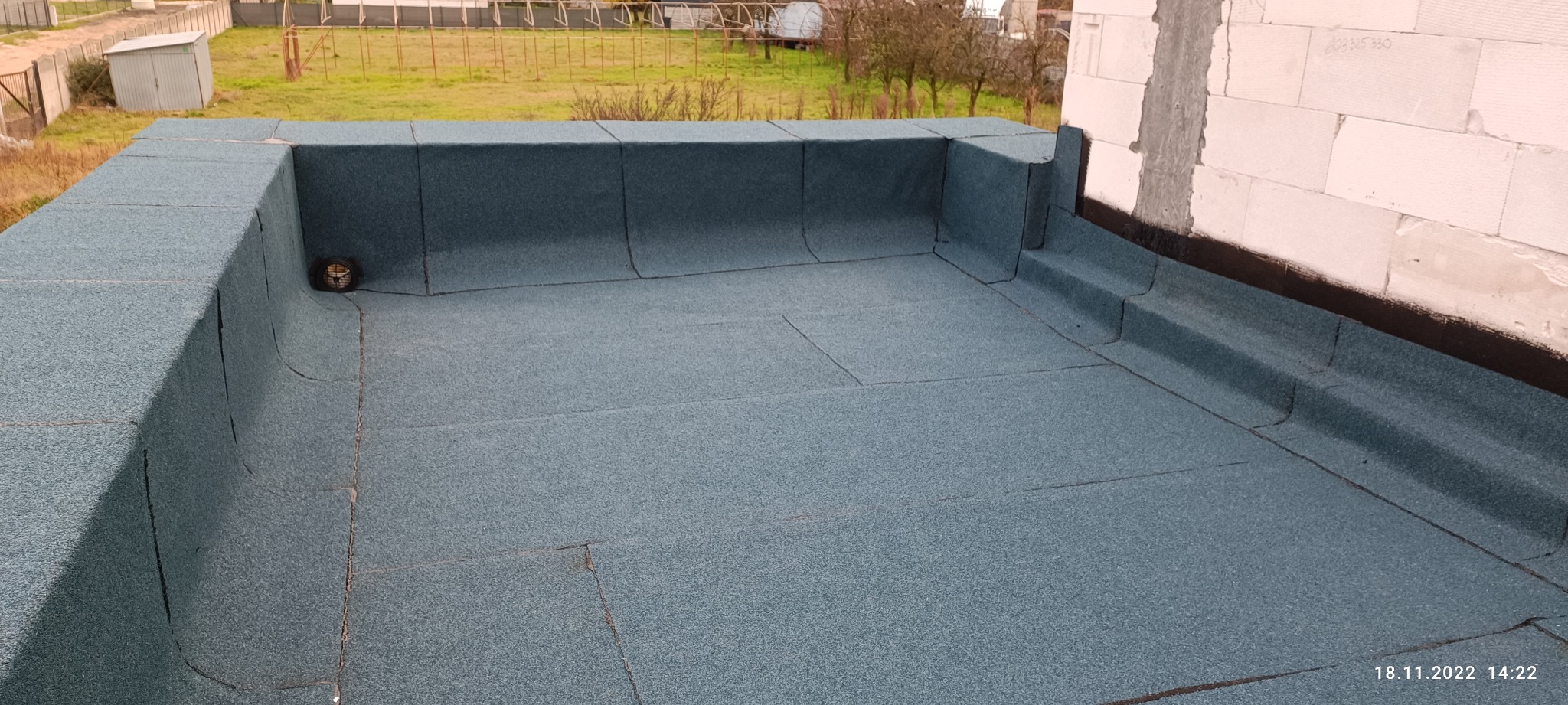 Wykończony płaski dach pokryty papą termozgrzewalną w kolorze ciemnego turkusu, widoczny kominek wentylacyjny i mur z betonu komórkowego w tle.