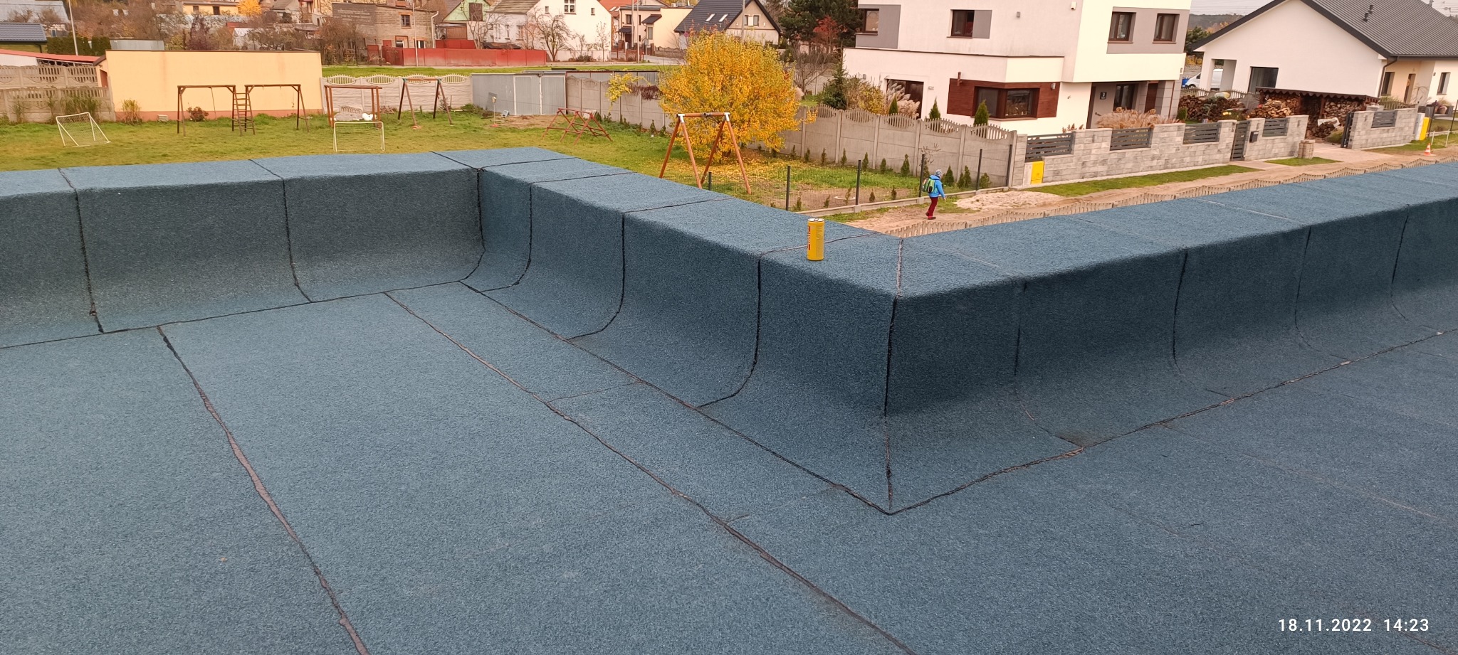 Widok z góry na płaski dach pokryty papą termozgrzewalną z widocznymi łączeniami, w tle budynki mieszkalne, drzewo z żółtymi liśćmi oraz plac zabaw.
