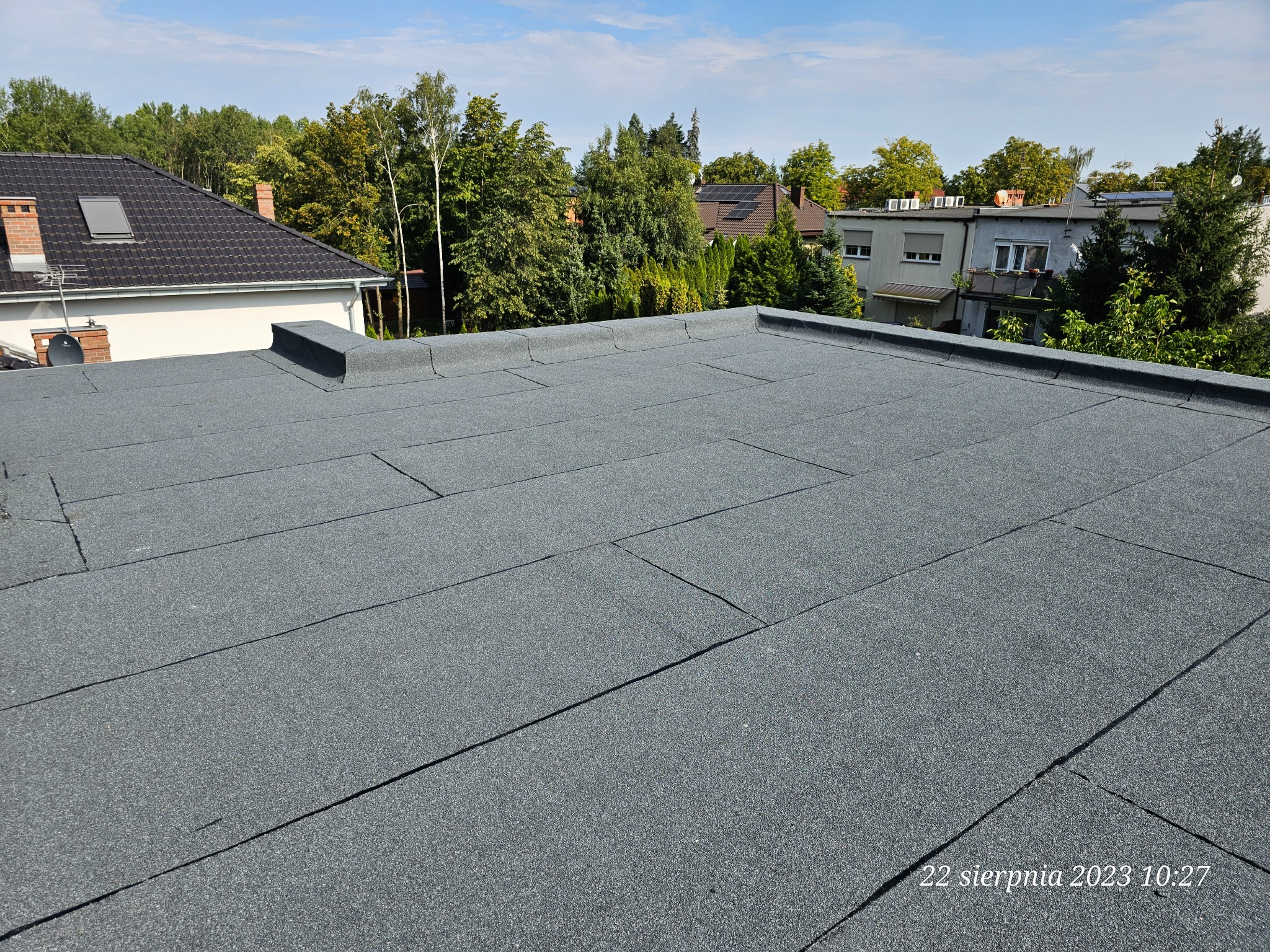Szczegółowe ujęcie płaskiego dachu pokrytego papą termozgrzewalną na tle zieleni drzew i zabudowań, wykonane w słoneczny dzień.