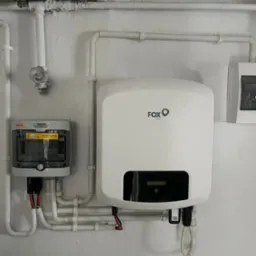 Biały falownik FoxESS zamontowany na ścianie, widoczne kable i przewody elektryczne, obok skrzynka zabezpieczeń KENO, instalacja w pomieszczeniu z rurami.