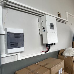 TEL-CARS Andrzej Pudło - Biała skrzynka rozdzielcza i falownik fotowoltaiczny zamontowane na szarej ścianie, widoczne rury instalacyjne i kartony na podłodze.