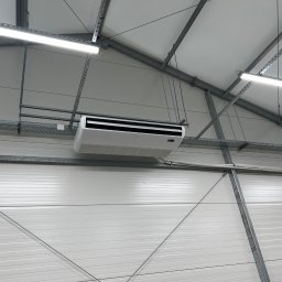 TEL-CARS Andrzej Pudło - Biała jednostka klimatyzacyjna zamontowana pod sufitem w hali przemysłowej, widoczne stalowe elementy konstrukcyjne i oświetlenie LED.