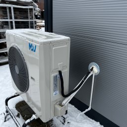 TEL-CARS Andrzej Pudło - Zewnętrzna jednostka klimatyzacji z niebieskim logo na metalowym stelażu, częściowo zasypana śniegiem, przy szarej ścianie budynku z widocznymi przewodami wchodzącymi do wnętrza przez otwór...