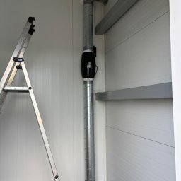 TEL-CARS Andrzej Pudło - Szczegół instalacji wentylacyjnej z czarnym wentylatorem kanałowym i spiralnymi rurami w pomieszczeniu o białych ścianach panelowych, widoczna drabina aluminiowa.