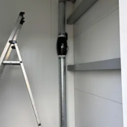 Szczegół instalacji wentylacyjnej z czarnym wentylatorem kanałowym i spiralnymi rurami w pomieszczeniu o białych ścianach panelowych, widoczna drabina aluminiowa.