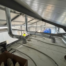 TEL-CARS Andrzej Pudło - Instalacja wentylacyjna z rurami spiro w hali przemysłowej, widok z góry na montaż, pracownik w żółtej kamizelce w tle, narzędzia i kable na podłodze.