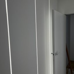 TEL-CARS Andrzej Pudło - Białe panele ścienne z wbudowanym oświetleniem LED i białe drzwi wewnętrzne z minimalistyczną klamką, widoczna drabina oparta o ścianę w tle.