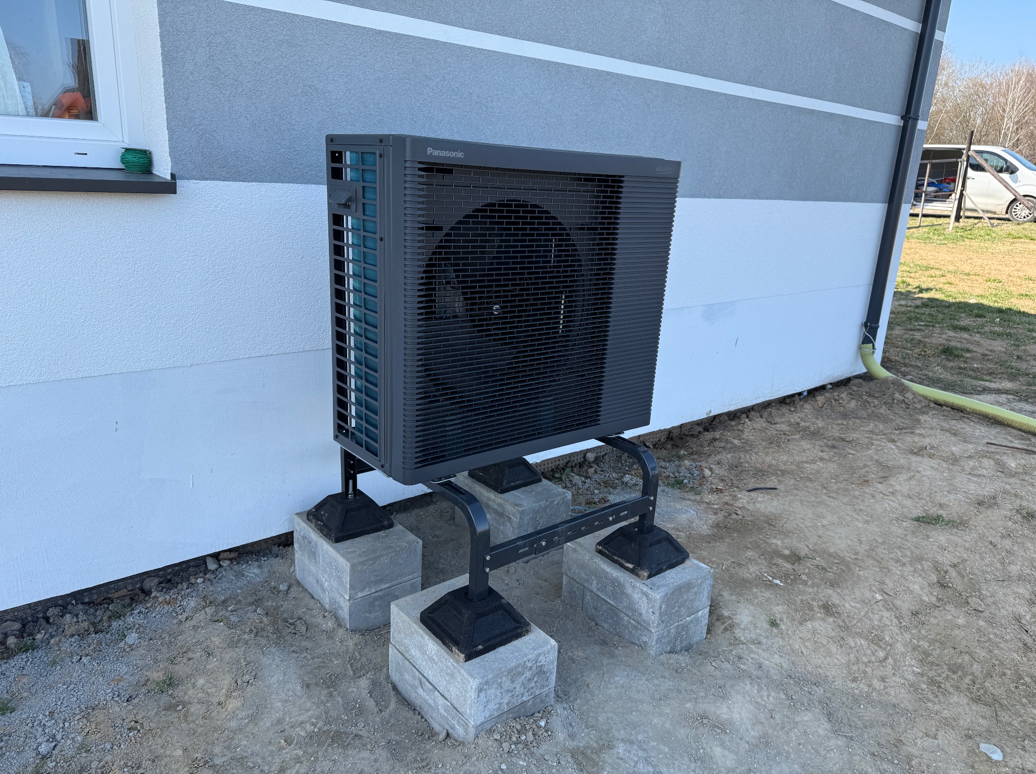 Zewnętrzna jednostka pompy ciepła Panasonic Aquarea zamontowana na betonowych bloczkach przy ścianie domu z szarym i białym tynkiem.