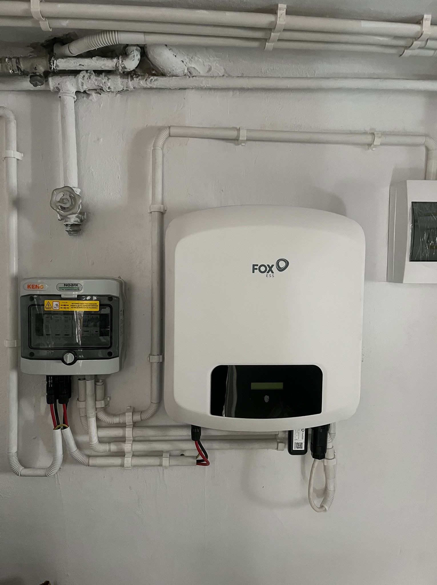Biały falownik FoxESS zamontowany na ścianie, widoczne kable i przewody elektryczne, obok skrzynka zabezpieczeń KENO, instalacja w pomieszczeniu z rurami.