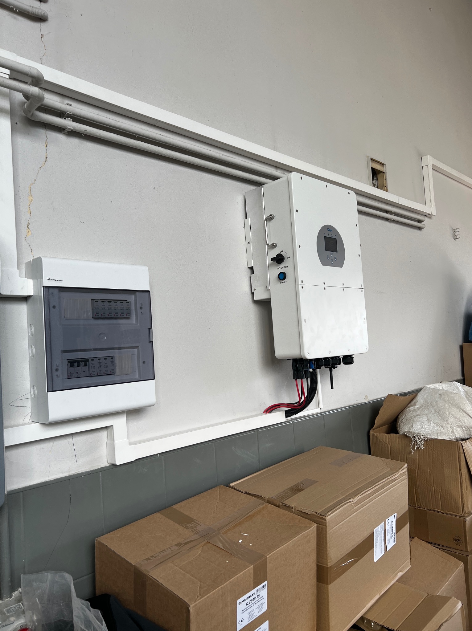Biała skrzynka rozdzielcza i falownik fotowoltaiczny zamontowane na szarej ścianie, widoczne rury instalacyjne i kartony na podłodze.