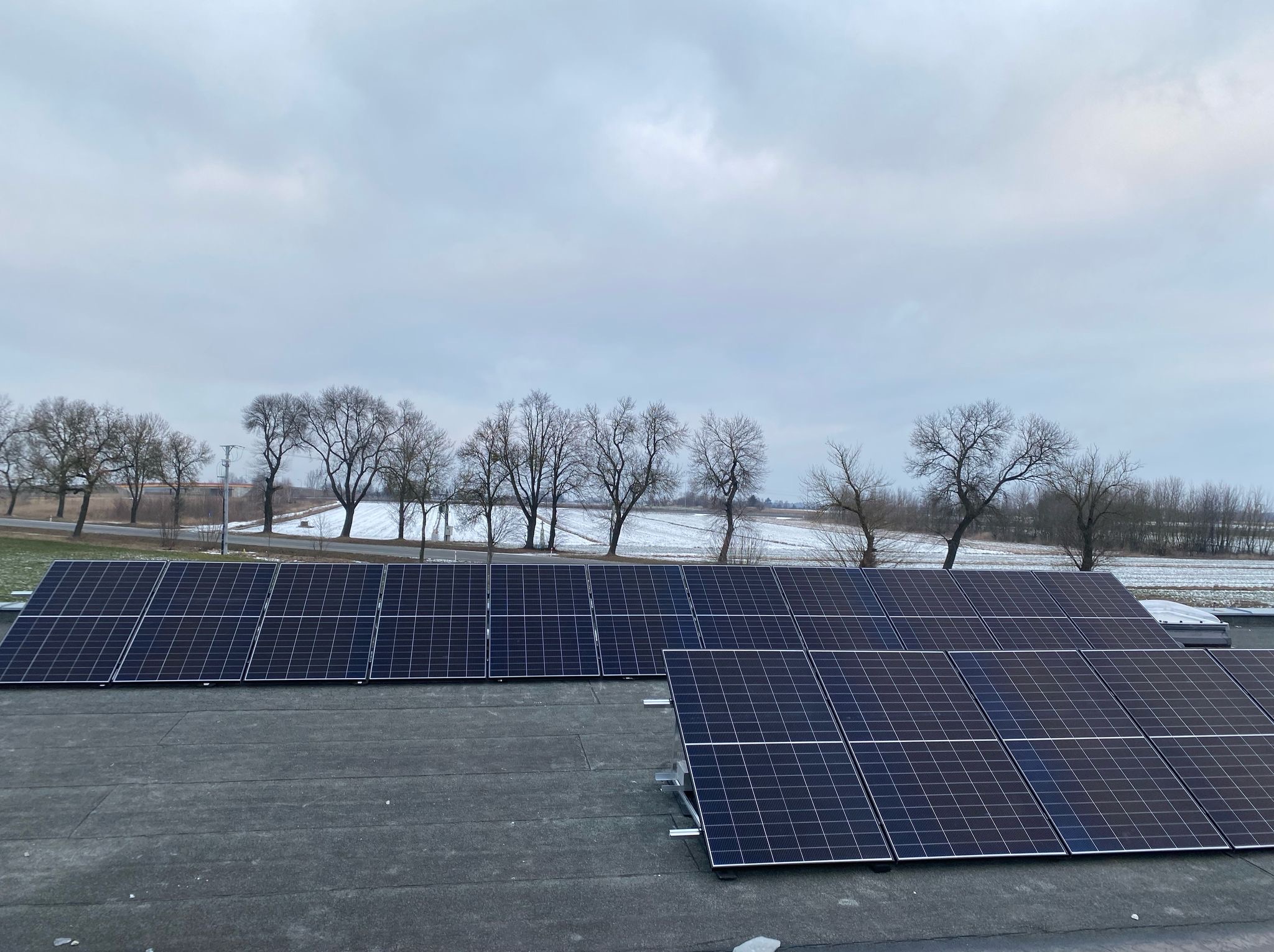 Panele fotowoltaiczne zamontowane na płaskim dachu z widokiem na zimowy krajobraz z polami i drzewami bez liści.