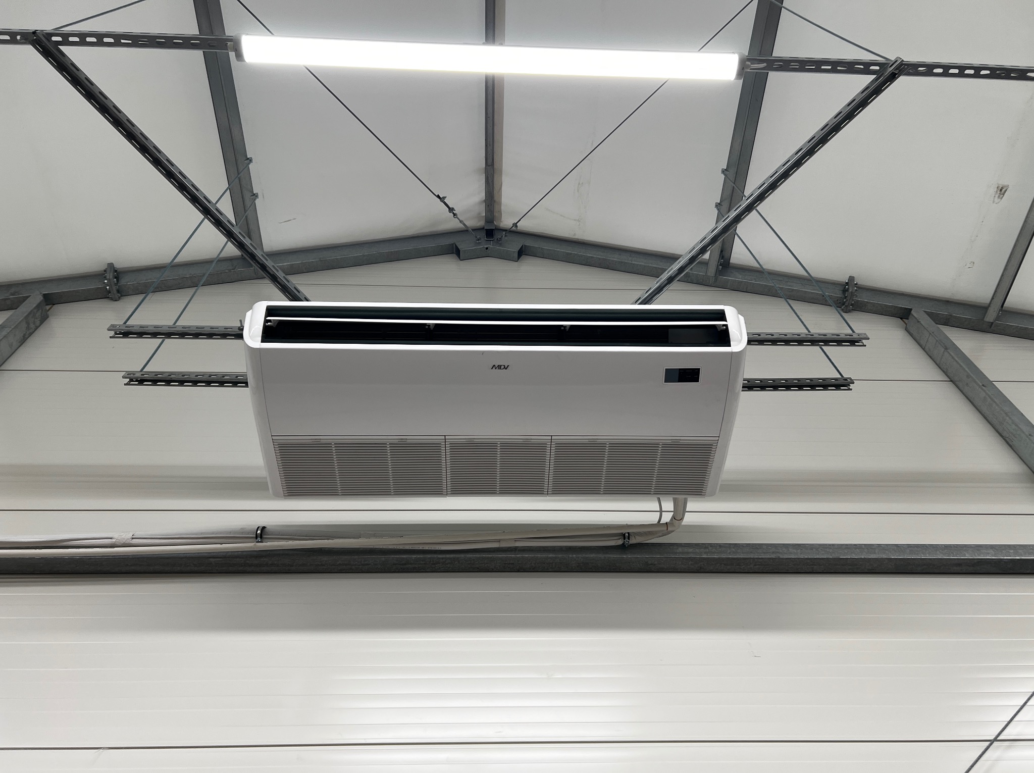 Biała jednostka klimatyzacyjna zamontowana pod sufitem hali produkcyjnej, widoczne stalowe profile konstrukcyjne i oświetlenie LED.