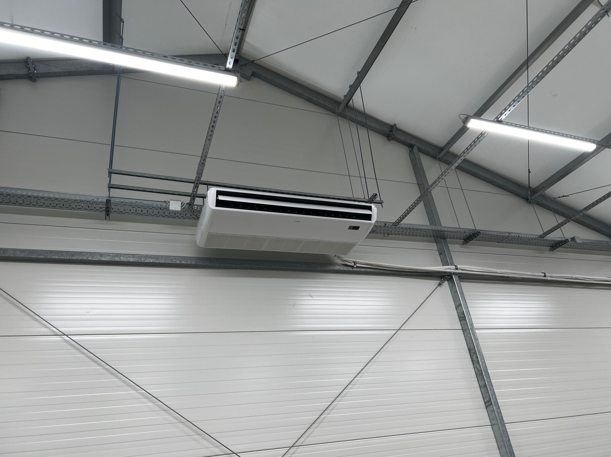 Biała jednostka klimatyzacyjna zamontowana pod sufitem w hali przemysłowej, widoczne stalowe elementy konstrukcyjne i oświetlenie LED.