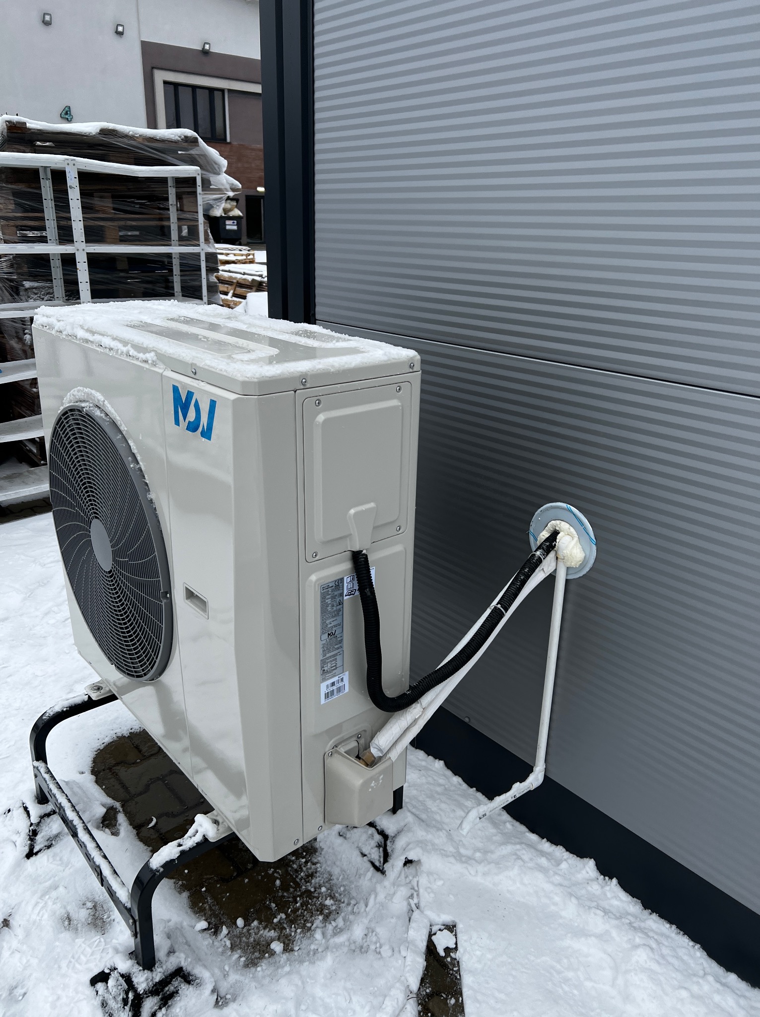 Zewnętrzna jednostka klimatyzacji z niebieskim logo na metalowym stelażu, częściowo zasypana śniegiem, przy szarej ścianie budynku z widocznymi przewodami wchodzącymi do wnętrza przez otwór...