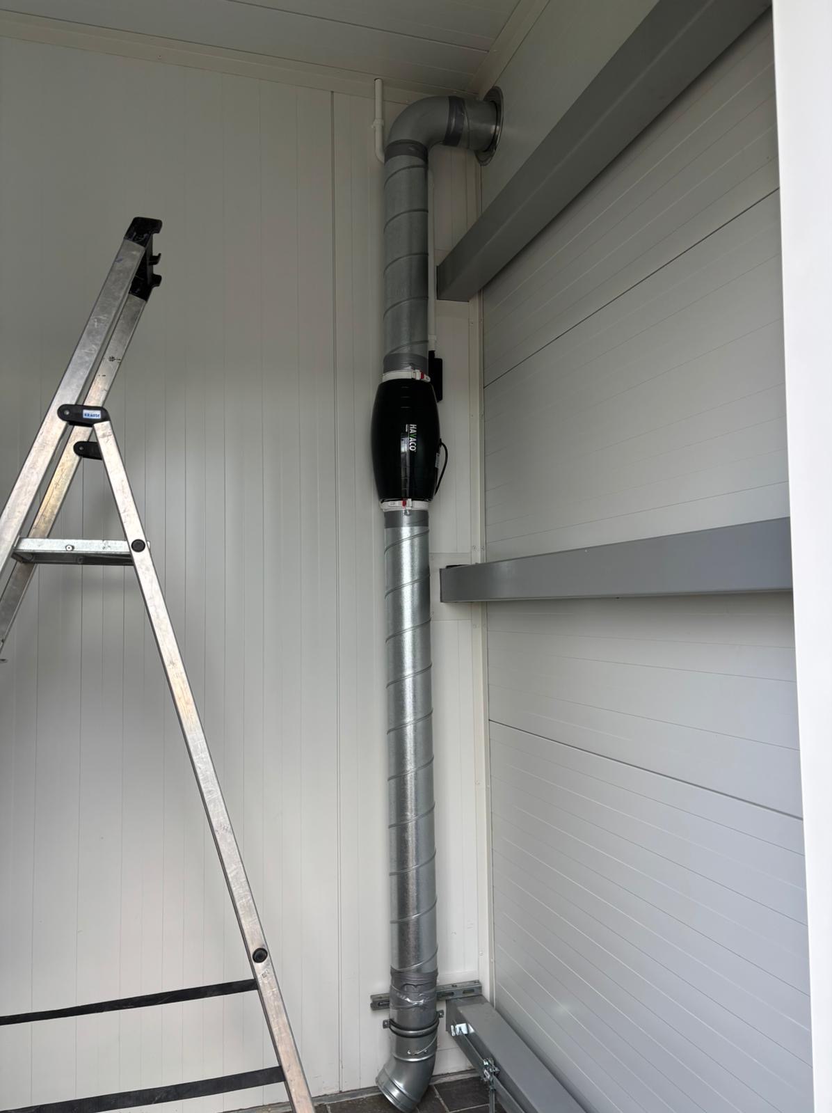 Szczegół instalacji wentylacyjnej z czarnym wentylatorem kanałowym i spiralnymi rurami w pomieszczeniu o białych ścianach panelowych, widoczna drabina aluminiowa.