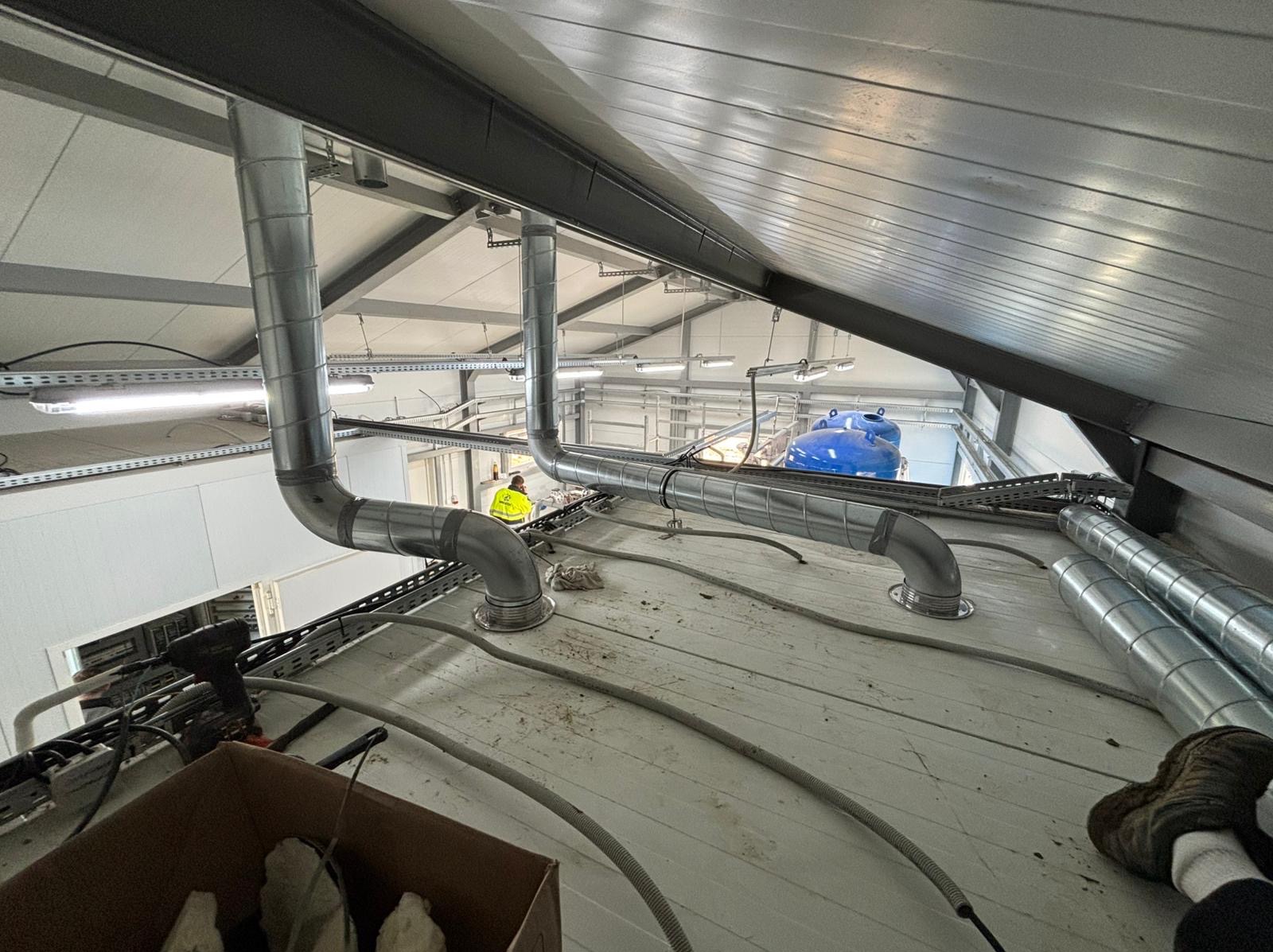 Instalacja wentylacyjna z rurami spiro w hali przemysłowej, widok z góry na montaż, pracownik w żółtej kamizelce w tle, narzędzia i kable na podłodze.