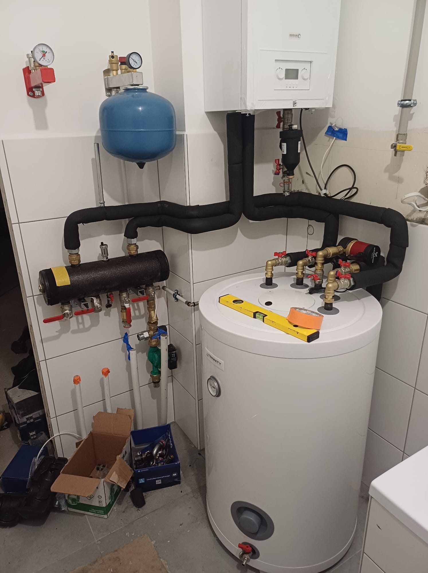 Kotłownia w trakcie montażu: biały zasobnik ciepłej wody Trinity z przyłączonymi rurami w izolacji, poziomica i taśma malarska na górze zasobnika, widoczne elementy instalacji grzewczej na ścianie...