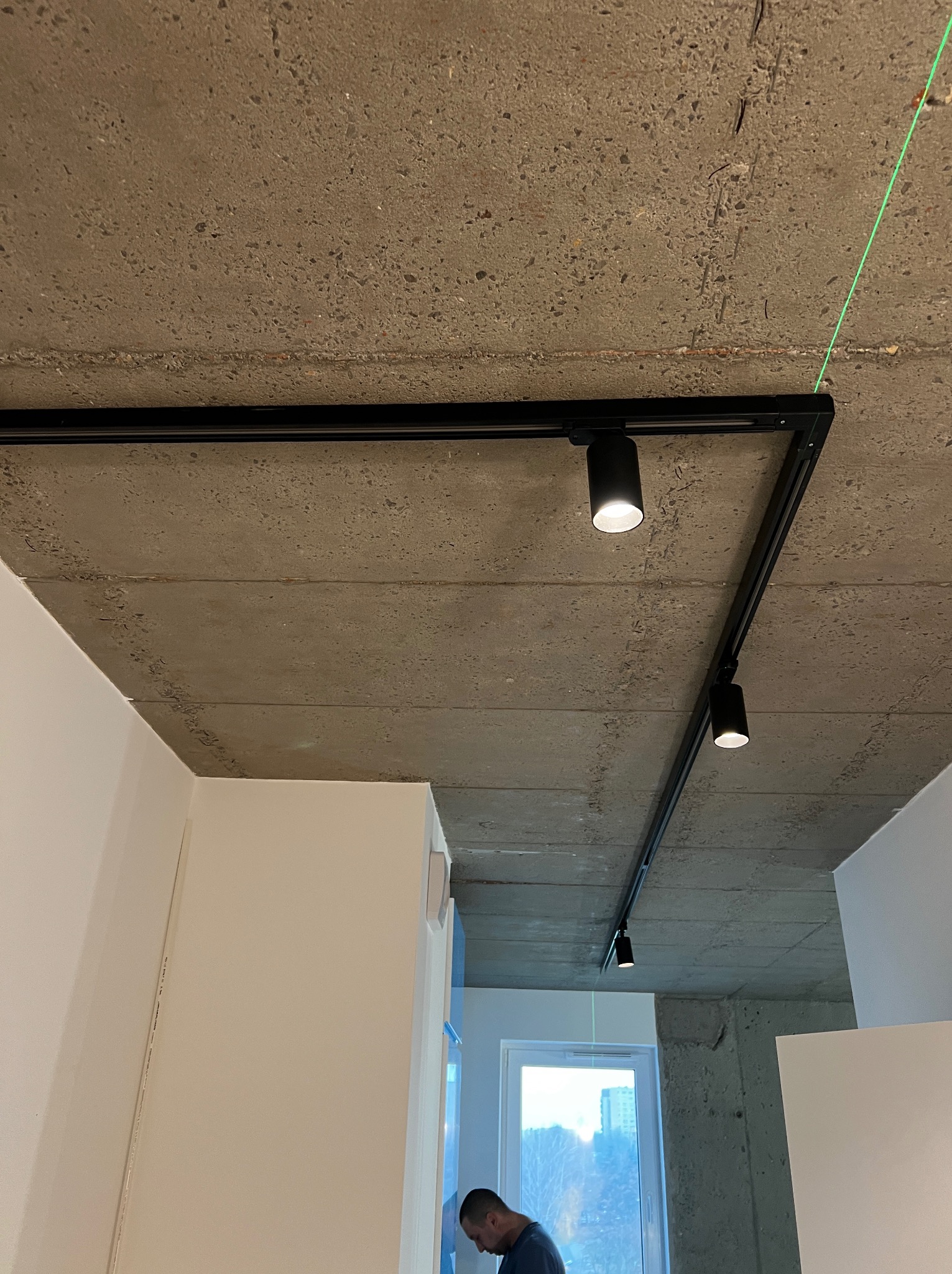 Czarna szyna oświetleniowa z reflektorami zamontowana na surowym betonowym suficie w Dęblinie, widoczny zielony laser wyznaczający linię prostą.