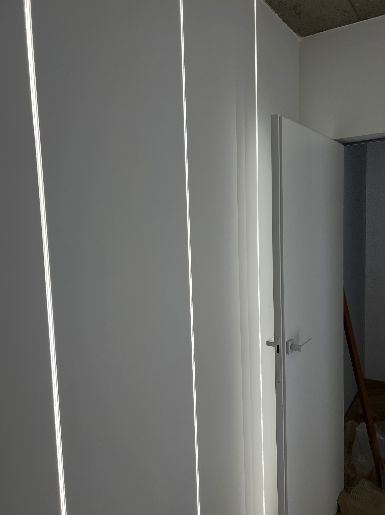 Białe panele ścienne z wbudowanym oświetleniem LED i białe drzwi wewnętrzne z minimalistyczną klamką, widoczna drabina oparta o ścianę w tle.