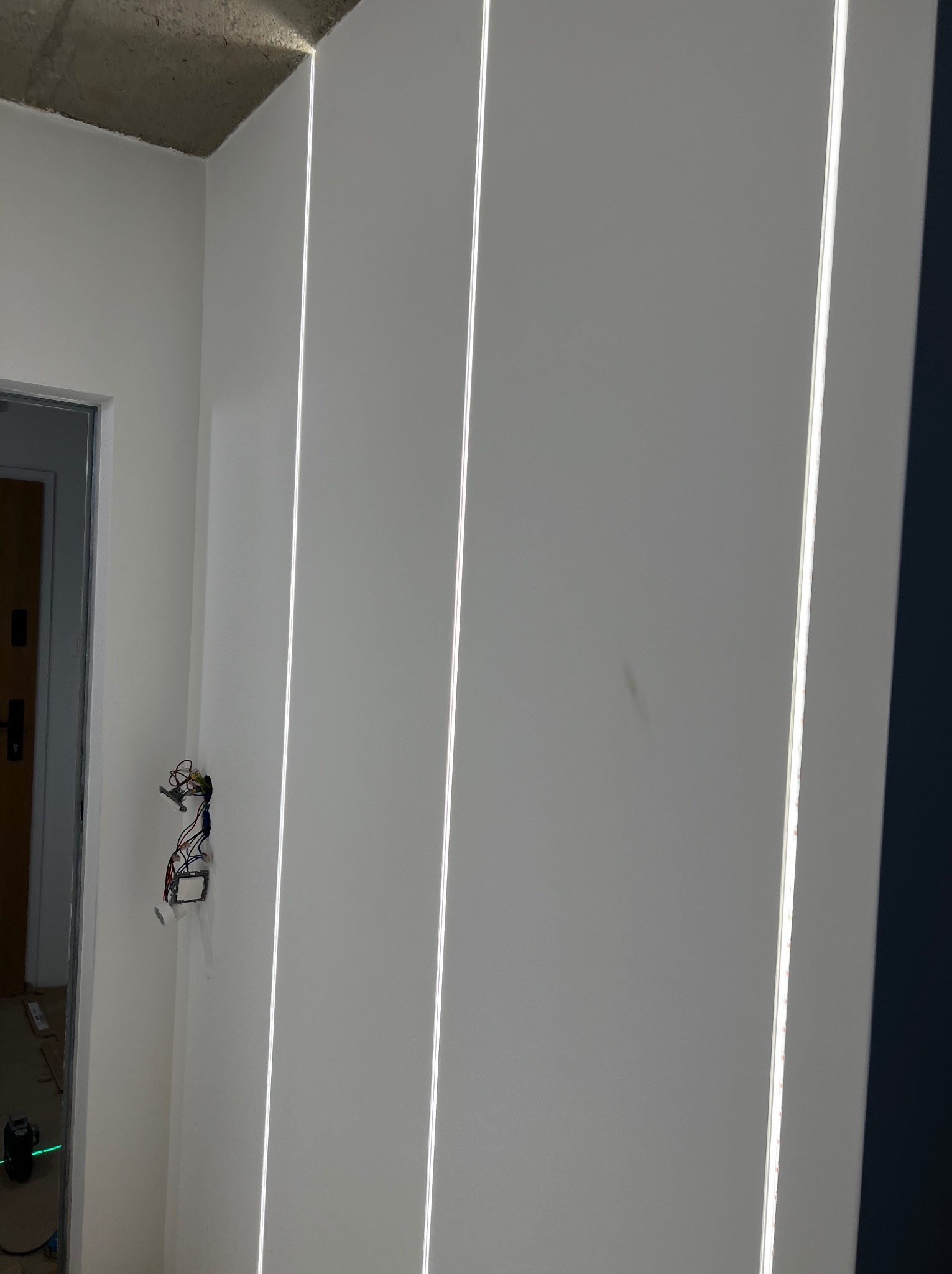 Wnętrze w trakcie remontu: pionowe, wpuszczane oświetlenie LED na białej ścianie, widoczne przewody elektryczne wystające ze ściany obok drzwi.