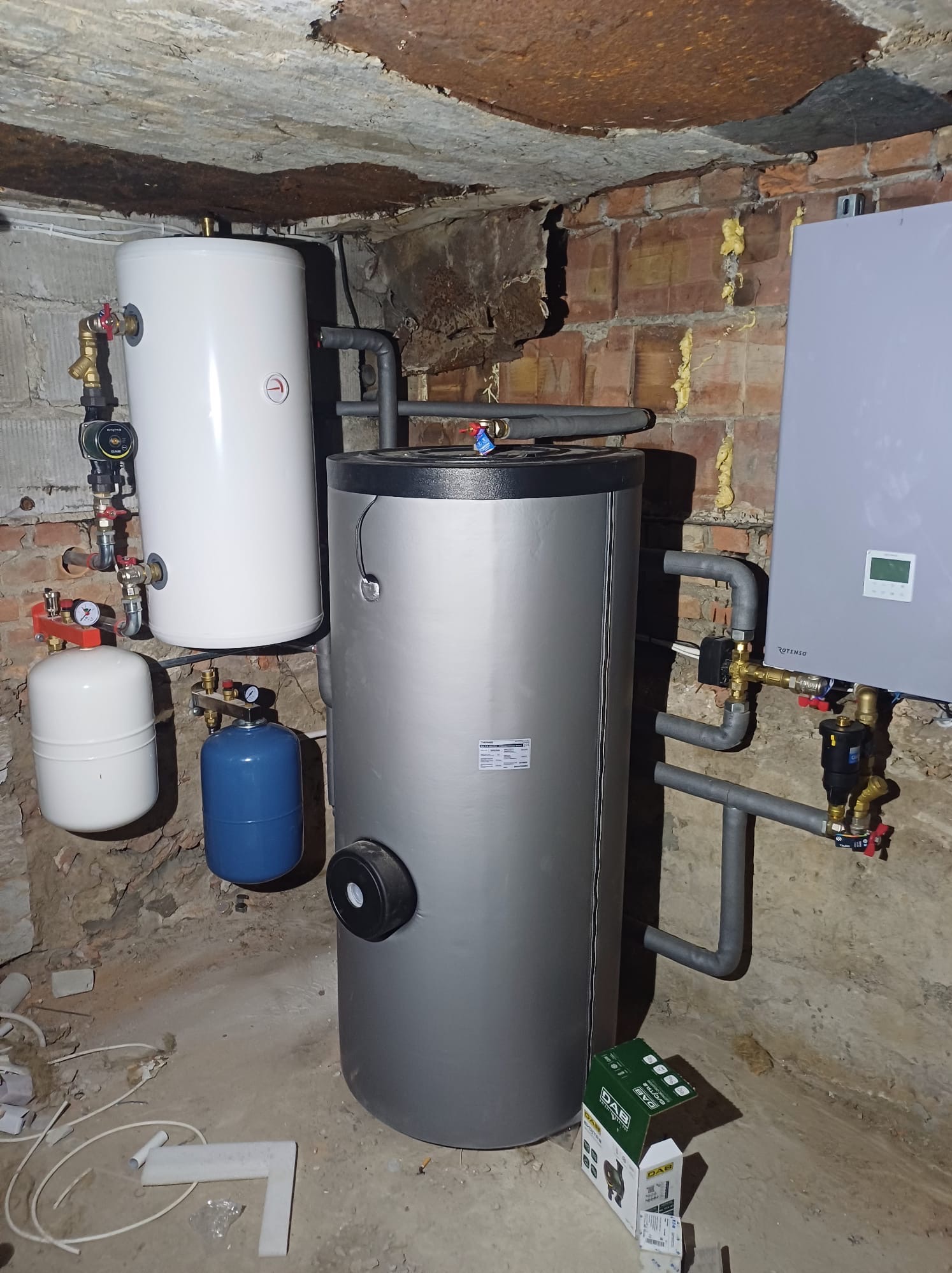 Instalacja grzewcza w piwnicy: zbiorniki akumulacyjne, naczynia przeponowe, kotłownia gazowa Rotenso, widoczne rury, zawory i manometry, surowe ceglane ściany.