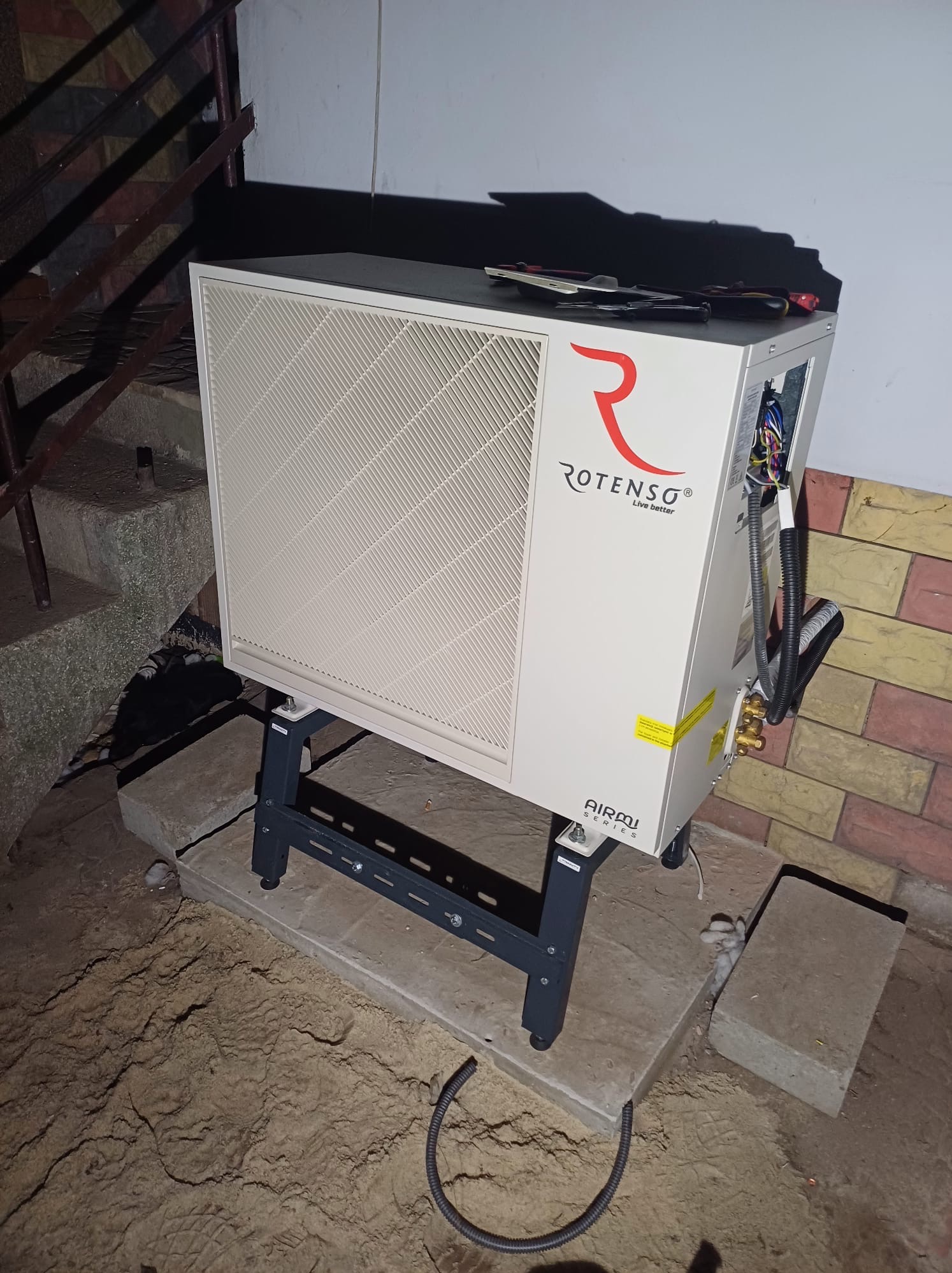 Zewnętrzna jednostka pompy ciepła marki Rotenso, model AIRMI, zamontowana na betonowych podkładach, z widocznymi przewodami i narzędziami na górze, w trakcie instalacji przy ścianie budynku...