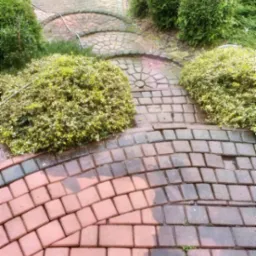 Częściowo umyta, mokra kostka brukowa w odcieniach czerwieni i szarości, otoczona zielonymi krzewami i drzewami, z widocznym wężem ogrodowym.