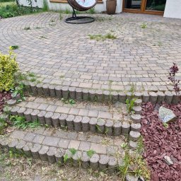 Sebo Dach I Bruk jak nowy - Zarośnięty, okrągły taras z kostki brukowej z schodami i wiszącym fotelem. Widoczne chwasty i mech między kostkami. Tło stanowi biały dom z brązowymi oknami.