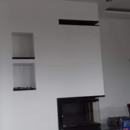 Biała ściana z nowoczesnym kominkiem obudowanym czarnym kamieniem, z wnękami i podwieszanym sufitem z oświetleniem punktowym.