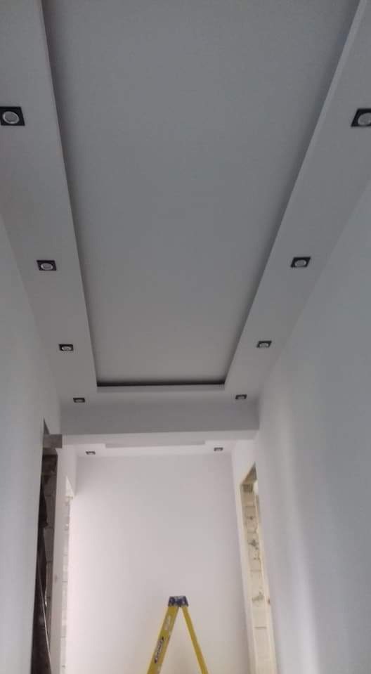 Białe wnętrze z podwieszanym sufitem z wbudowanym oświetleniem punktowym, widoczna żółta drabina w tle, ściany świeżo pomalowane.