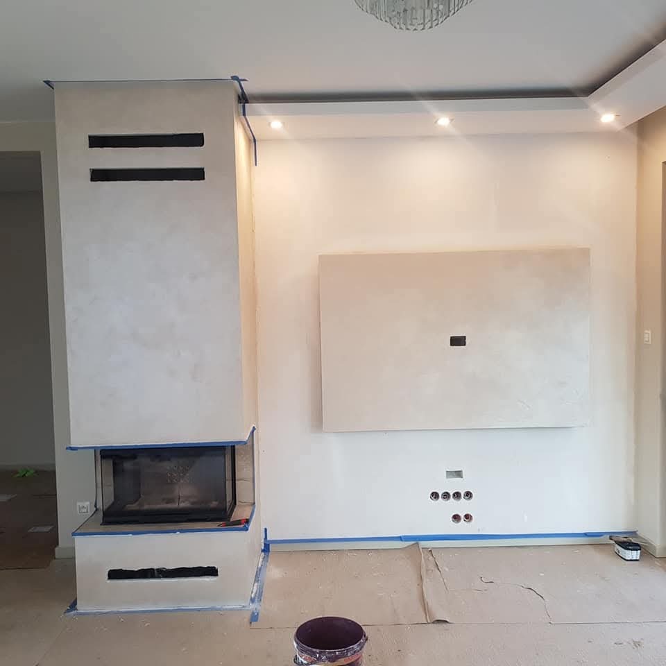 Ściana z kominkiem i przygotowanym miejscem na telewizor w trakcie prac wykończeniowych, zabezpieczona taśmą malarską i kartonem, widoczny otwarty pojemnik z farbą.