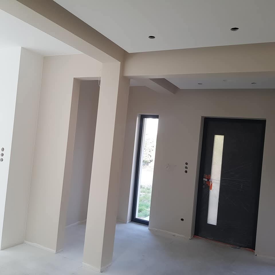 Wnętrze w trakcie prac wykończeniowych, ściany pomalowane na jasny beż, widoczna czarna, częściowo ofoliowana, nowa stolarka drzwiowa z przeszkleniem, surowy beton na podłodze.