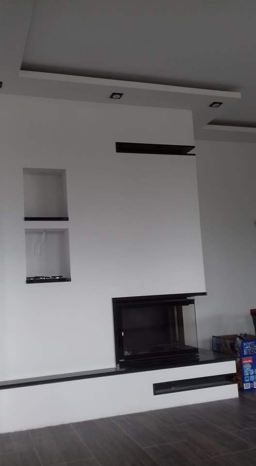 Biała ściana z nowoczesnym kominkiem obudowanym czarnym kamieniem, z wnękami i podwieszanym sufitem z oświetleniem punktowym.