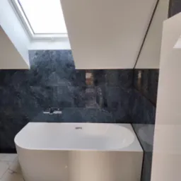 Nowoczesna łazienka na poddaszu z białą wanną, marmurową podłogą i ciemnozielonymi płytkami na ścianie. Okno dachowe wpuszcza naturalne światło.