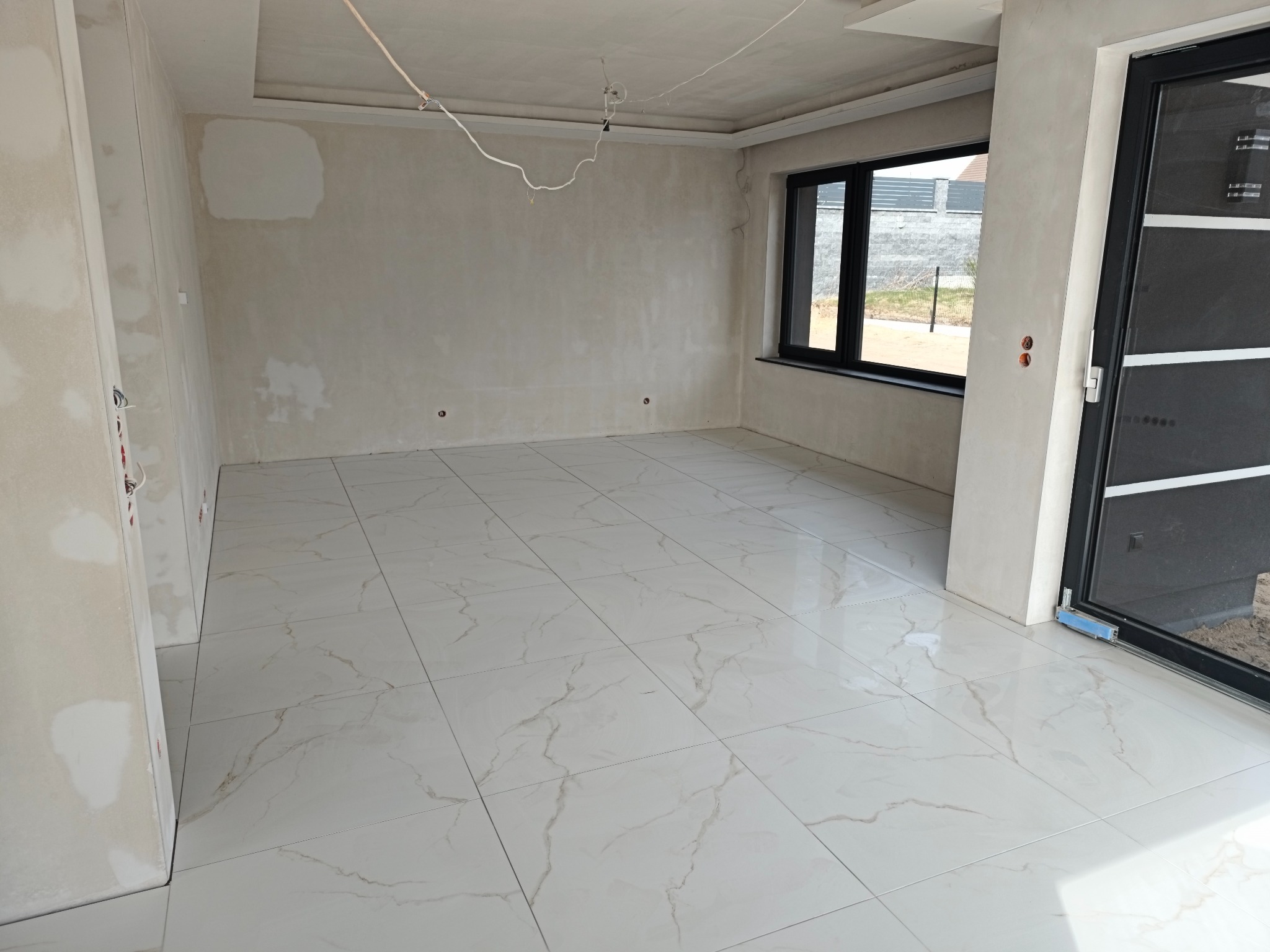 Wnętrze w stanie deweloperskim z wykończonymi podłogami z płytek imitujących marmur. Ściany przygotowane pod malowanie, widoczne okna i drzwi balkonowe.