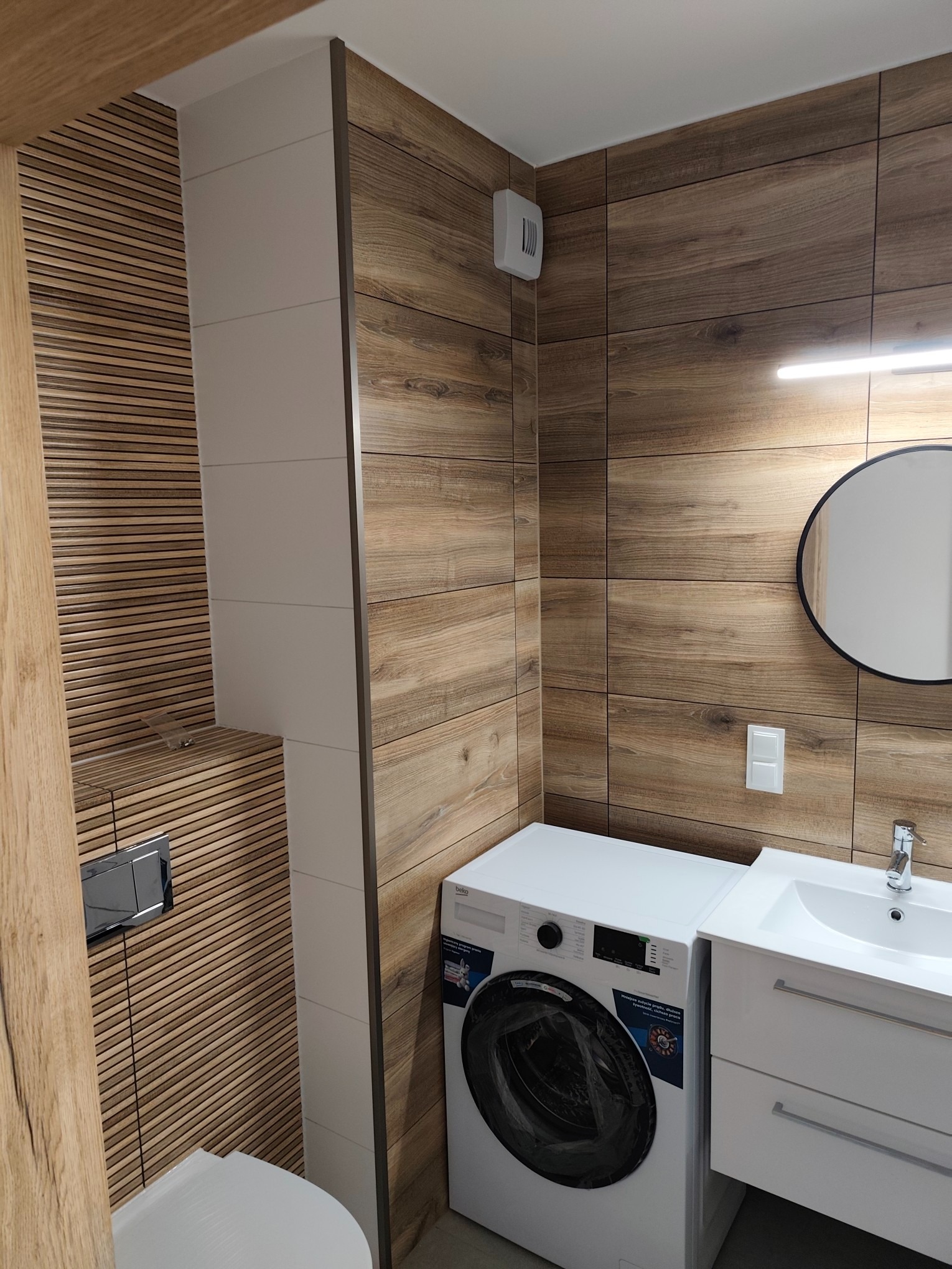 Nowoczesna łazienka z drewnopodobnymi płytkami, białą armaturą i pralką. Umywalka z szafką, okrągłe lustro. Minimalistyczny design, ciepła kolorystyka.