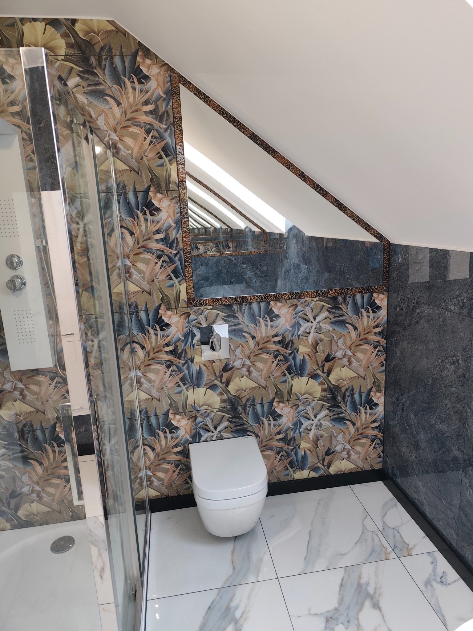 Nowoczesna łazienka na poddaszu z kabiną prysznicową, toaletą i marmurową podłogą. Ściany wyłożone płytkami z motywem roślinnym i ciemnymi płytkami imitującymi kamień.