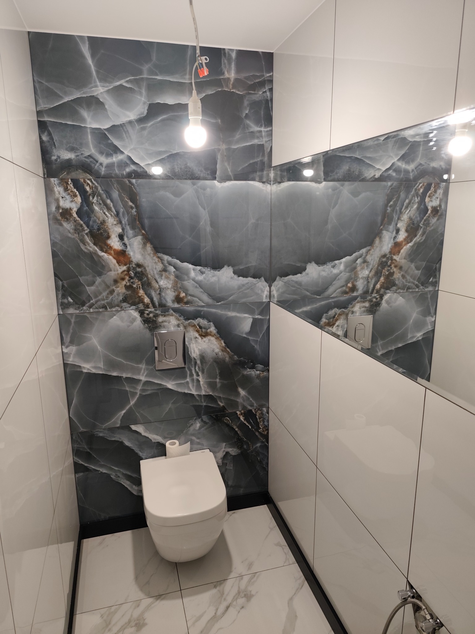 Nowoczesna toaleta z marmurowymi płytkami w odcieniach szarości i bieli, podświetlona żarówką, z widocznym fragmentem lustra i minimalistyczną armaturą.
