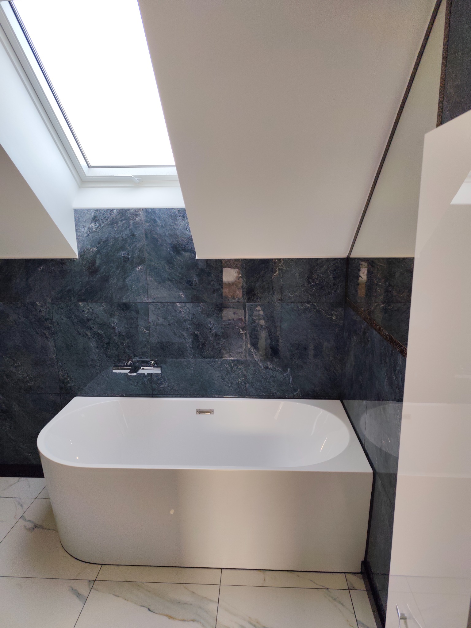 Nowoczesna łazienka na poddaszu z białą wanną, marmurową podłogą i ciemnozielonymi płytkami na ścianie. Okno dachowe wpuszcza naturalne światło.