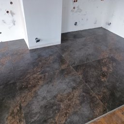 MATS-BUD - Ułożone ciemne płytki podłogowe imitujące kamień w surowym wnętrzu z widocznymi puszkami elektrycznymi i śladami po gładzi na ścianach.