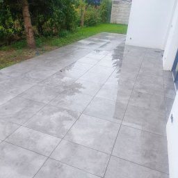 MATS-BUD - Nowoczesny taras z szarych, kwadratowych płytek, częściowo mokrych po deszczu, przylegający do białej ściany budynku i zielonego trawnika.