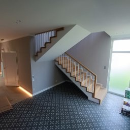 MATS-BUD - Nowoczesne wnętrze po remoncie: szare płytki podłogowe we wzory, drewniane schody z białymi balustradami, listwy przypodłogowe z oświetleniem LED, paczki kleju do płytek przy oknie.