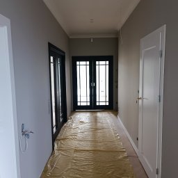 MATS-BUD - Korytarz w trakcie malowania ścian na szaro, podłoga zabezpieczona folią, widoczne czarne przeszklone drzwi i białe drzwi po prawej stronie, wystające kable ze ściany po lewej.